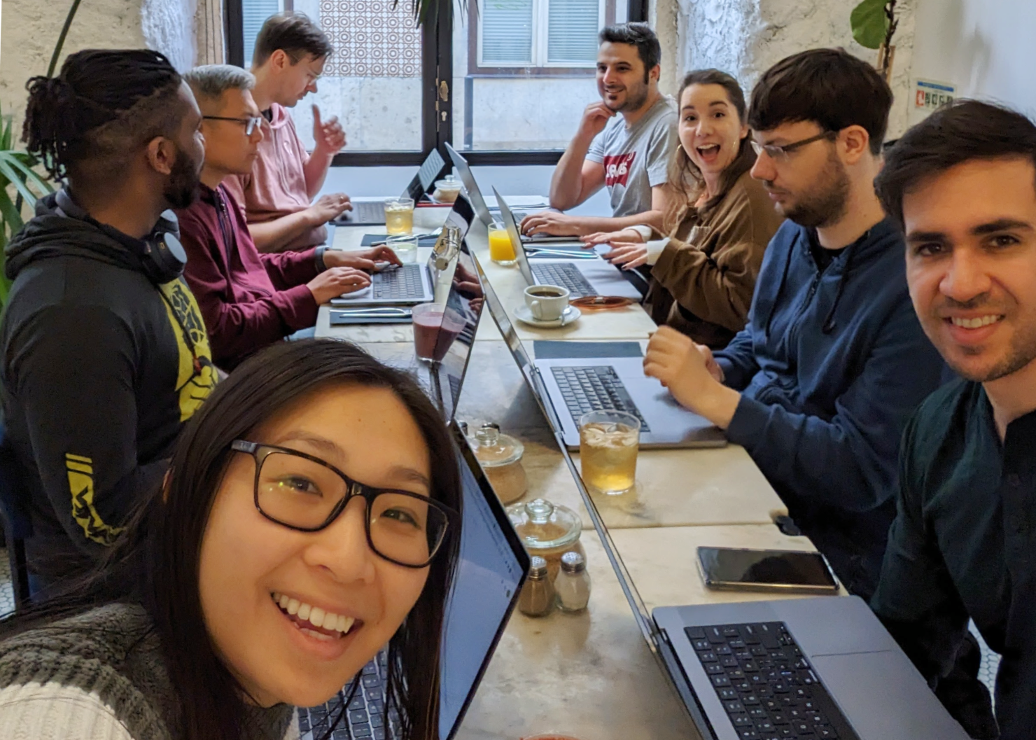 Jenny takes a selfie including the team all sitting at a long table in a room with stone and Portuguese tiled walls with laptops, coffees, and juices. David, Mateusz, Laura, Farzad, Anders, Kevin, and Gavin are all working, looking at the camera, or chatting to each other.